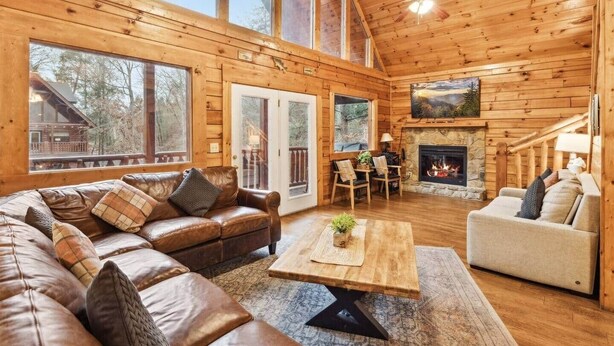 A wooden cabin interior with a leather sofa, a coffee table, a fireplace, and large windows.
