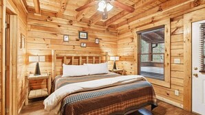 A wooden cabin bedroom with a bed, two bedside tables, a ceiling fan, and a window with blinds.