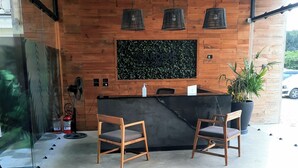 A reception area with wooden panel walls, a black reception desk, two wooden chairs, a large potted plant, and a wooden coffee table with magazines. - Beautiful & Fully Equipped Condo Studio Strategically located (Tulum)