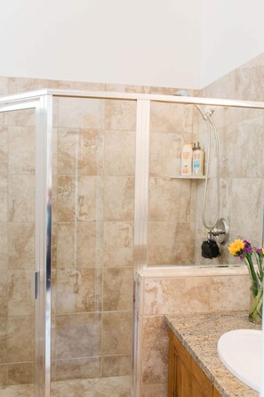 Un baño con cabina de ducha de vidrio, pared con azulejos de mármol, tocador de madera y un estante pequeño con artículos de aseo.