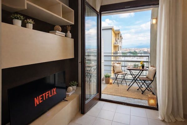 A modern living room with a flat-screen TV, a balcony with a table and chairs, and a city view.