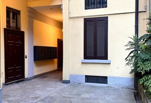 A narrow alleyway with a building featuring a dark wooden door, a window with shutters, and a small planter with green foliage.