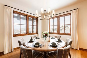 Uma sala de jantar com uma mesa redonda posta para seis pessoas, grandes janelas e um candelabro.