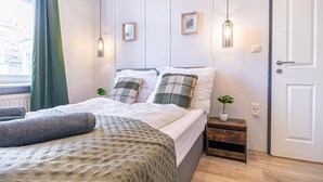 A neatly made bed with a patterned bedspread, a small bedside table with a plant, and a door to the right. - Bright & Modern Room in Vienna’s Meidling District (Vienna)