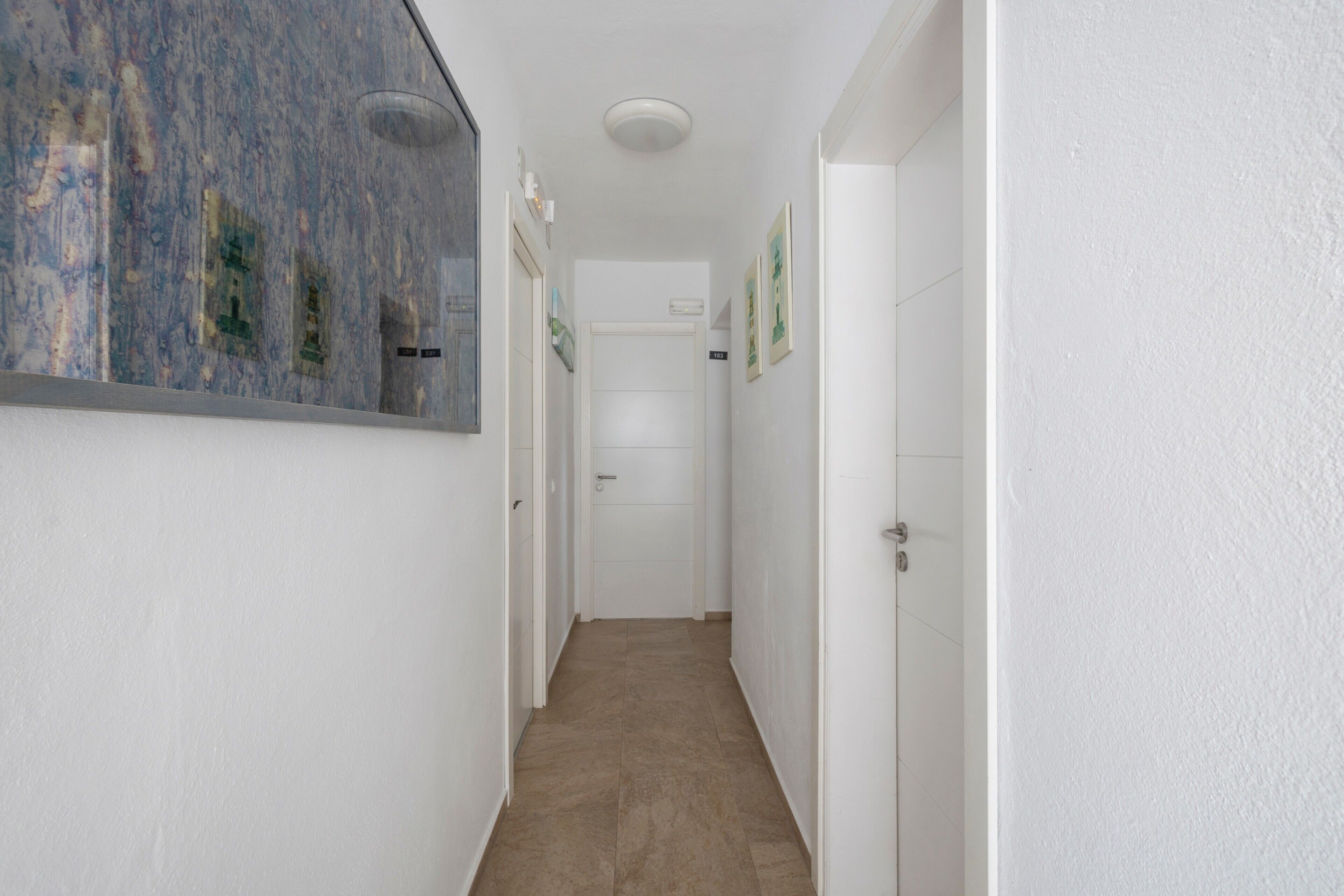 Un couloir étroit aux murs blancs, doté d’un sol carrelé et d’une porte au fond.