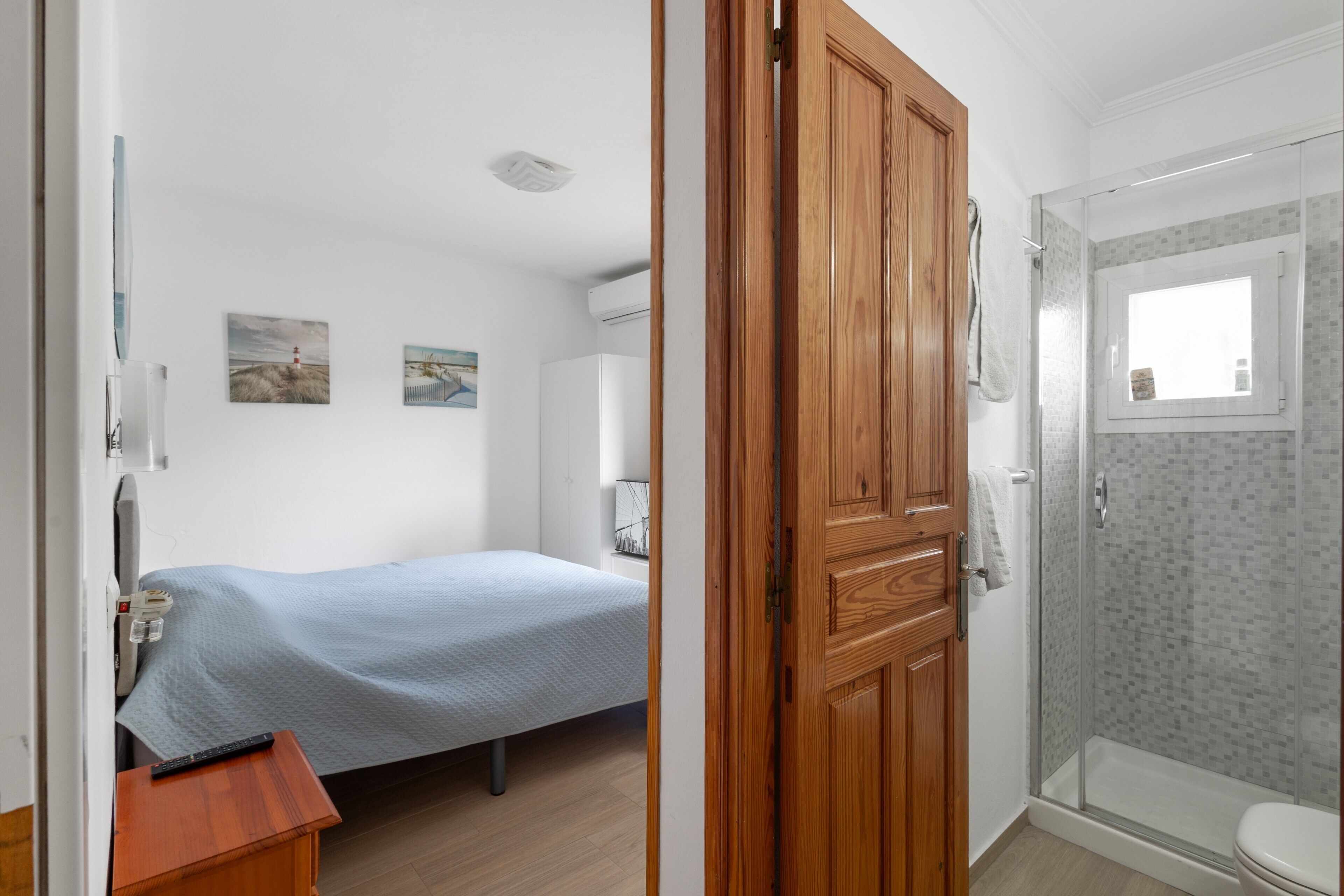 A bedroom with a bed, a wooden door, a shower area, and a small table with a remote control.