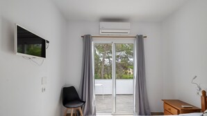 Une chambre avec un lit, un téléviseur fixé au mur, une chaise, une table de chevet en bois et une porte-fenêtre coulissante donnant sur un espace extérieur.