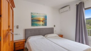 Quarto com cama, mesa de cabeceira, guarda-roupa de madeira, pintura de um pôr do sol, aparelho de ar condicionado e janela com cortinas.
