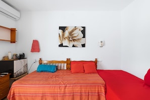 A bedroom with a bed, a wooden headboard, a red and orange striped bedspread, a red pillow, a blue throw blanket, a wall-mounted air conditioner, a shelf with a picture frame, and a small red object on the wall.