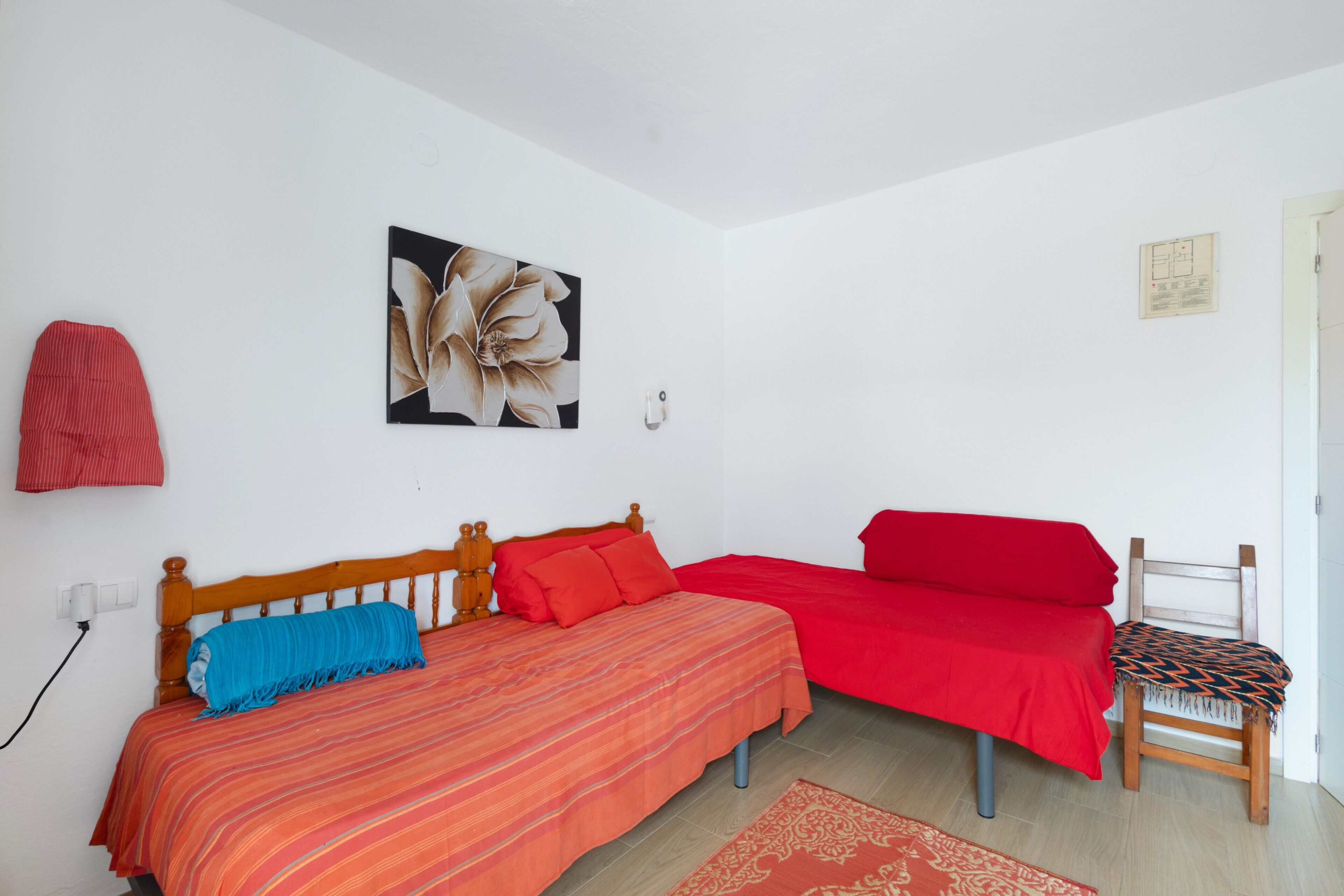 Una habitación con dos camas, una con una colcha a rayas y la otra con una colcha roja lisa. Hay una silla de madera con un cojín estampado, un interruptor de luz en la pared y un cuadro con motivos florales.