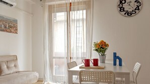 A room with a sofa, a dining table with chairs, a clock, and a window with sheer curtains. - Homiday - Hospital/Airport- A/C and Free Parking (Pisa)