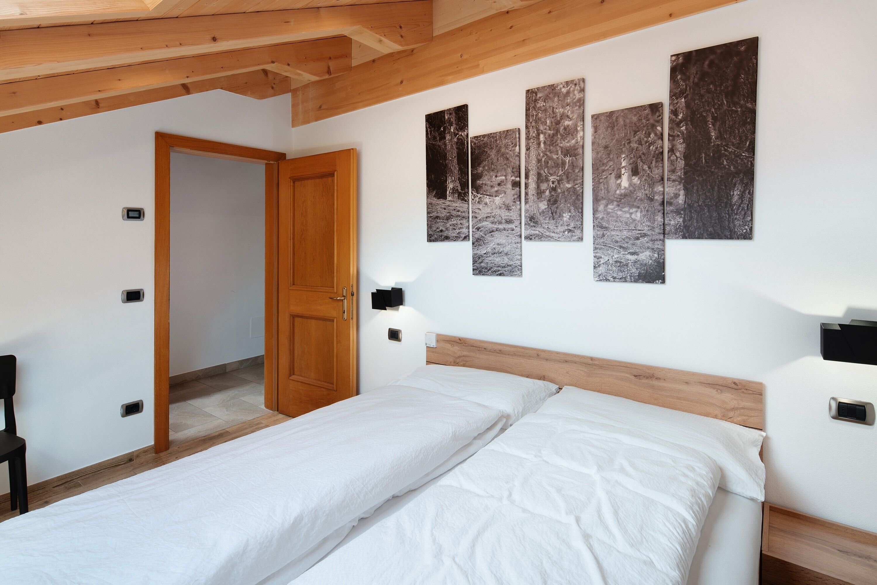 Un dormitorio con techo de madera, una cama con ropa blanca, una puerta de madera y tres fotografías blanco y negro en la pared.