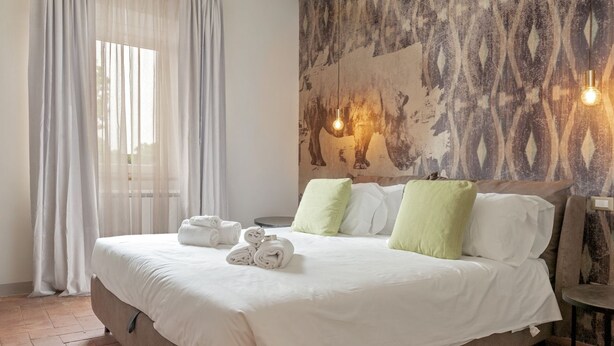 A neatly made bed with white linens and rolled towels, a patterned wall, and a hanging light fixture. - RINOCERONTE: Relax and enjoy this simple yet elegant apartment, equipped with all the comforts: PRIVATE parking, FULL HD TV, AIR CONDITIONING, and a fully equipped bathroom with a walk-in shower. All of this just 10 minutes from Pisa and 25 minutes from  (San Giuliano Terme)