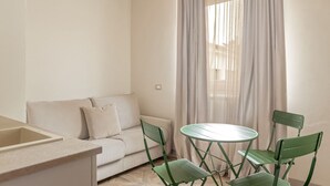 Una habitación pequeña y luminosa con un sofá, una mesa de comedor con sillas y una ventana con cortinas ligeras.