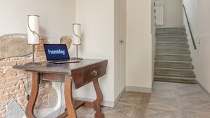 Una habitación con un escritorio de madera, una computadora portátil y dos lámparas. Se ve una escalera al fondo.
