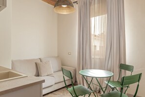 A small, well-lit room with a sofa, a dining table with chairs, and a window with sheer curtains.