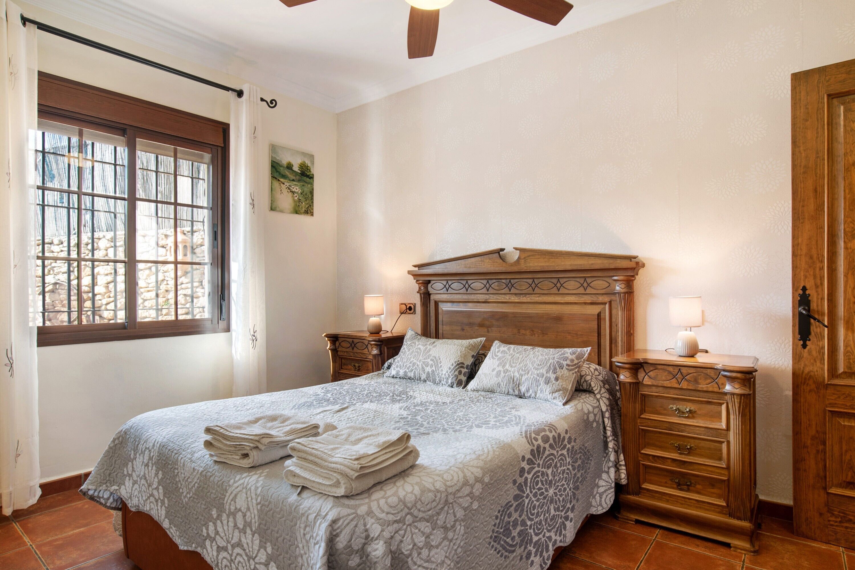 Un dormitorio con una cama grande, cabecera de madera, dos mesitas de noche con lámparas, una ventana con cortinas y un armario de madera.