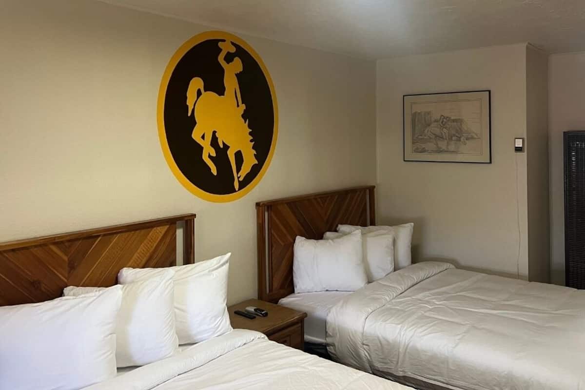 A hotel room with two beds, a wooden headboard, white bedding, a nightstand with a phone, a wall-mounted artwork, and a door with a window.