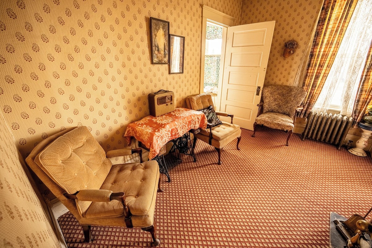 A vintage room with patterned wallpaper, a dining table with a floral tablecloth, a sofa, and a chair.