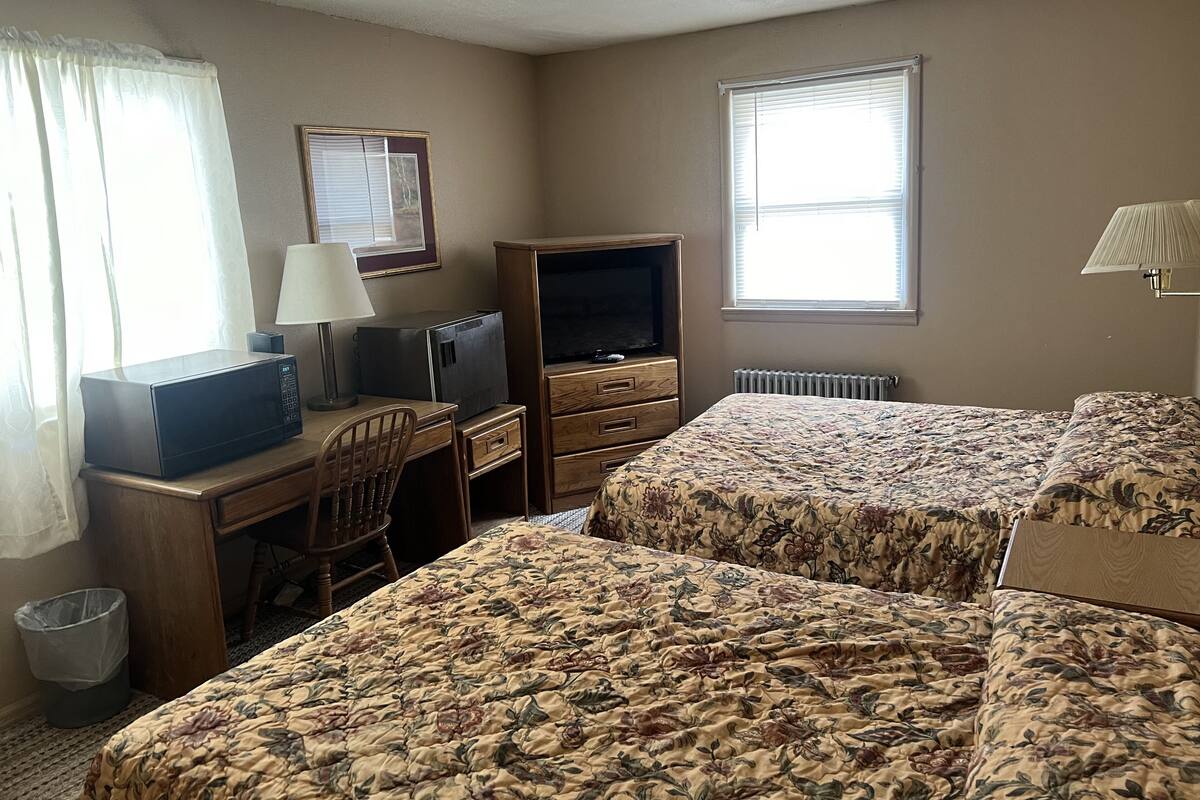 A hotel room with two beds, a desk with a television, a chair, a lamp, and a window with blinds.