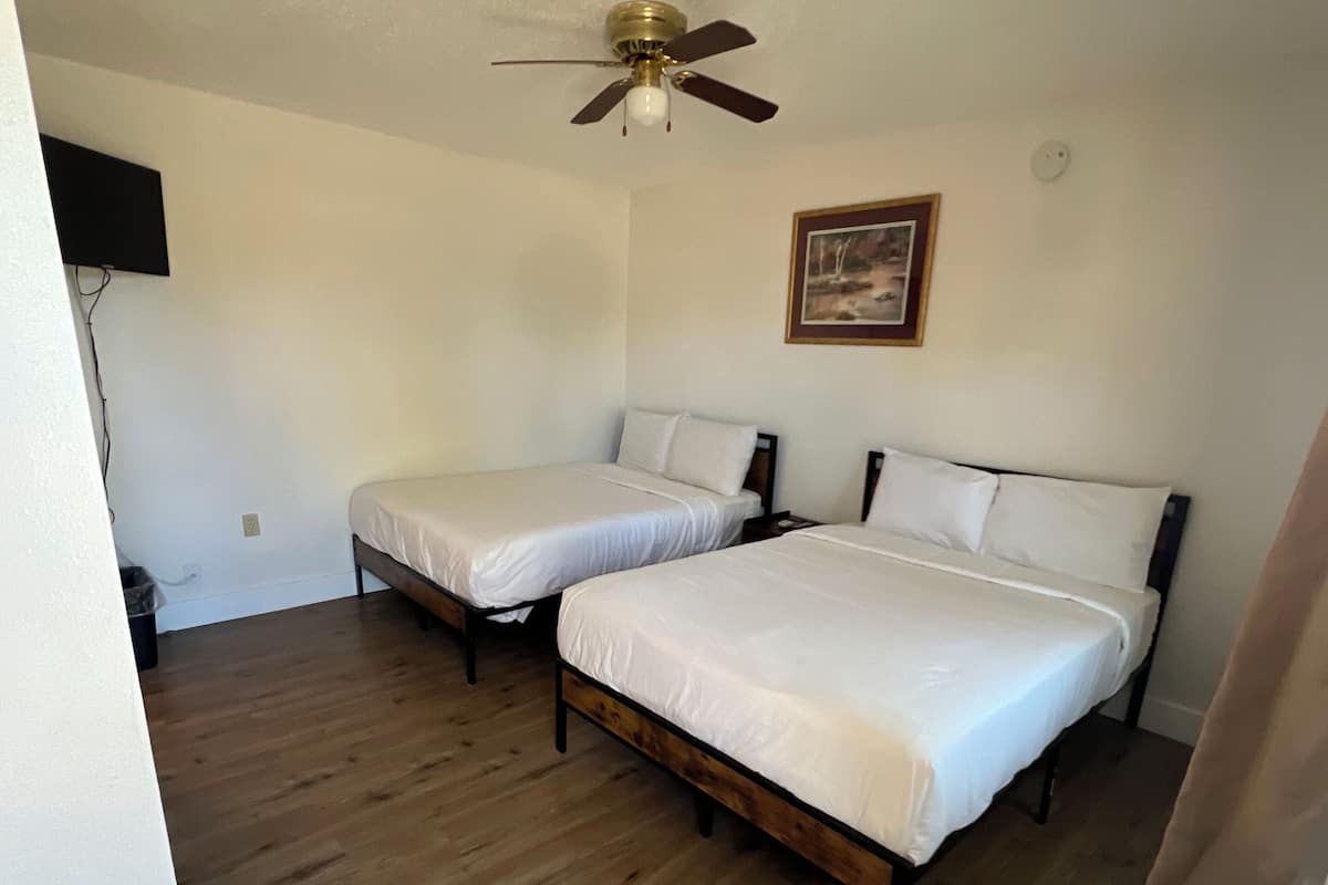 A room with two beds, a ceiling fan, a painting on the wall, and a wooden floor.