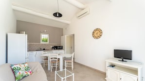 A compact living space with a kitchenette, a dining area, a sofa with a decorative pillow, a TV on a white cabinet, and a wall clock. - Holiday Home 'Chez Céline' with Private Pool, Wi-Fi and Air Conditioning (Aubignan)