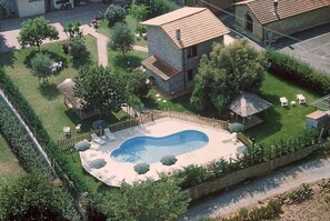 Ovenfra ses et boligområde med en swimmingpool, en indhegnet have og en lille bygning med et tag af fliser.