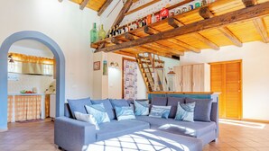 A cozy living room with a grey sofa, wooden ceiling beams, and a kitchen area visible through an archway. - La Casa Di Gio - Dépendance in Villa (Viagrande)
