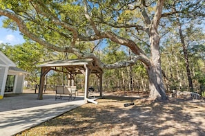 A covered outdoor seating area with a table and chairs, surrounded by trees.