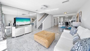 Una sala de estar moderna con un televisor grande, un sofá blanco, una mesa de centro de madera y una mesa de comedor de cristal.