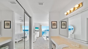 Habitación de hotel con un espejo grande, tocador con lavamanos, cama y balcón con vistas al océano.