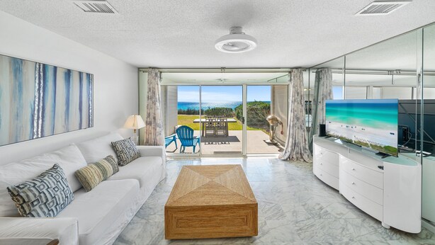 Una sala de estar moderna con un sofá blanco, una mesa de centro de madera y una pantalla grande mostrando una escena de playa.