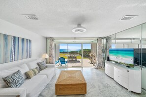 Una sala de estar moderna con un sofá blanco, una mesa de centro de madera y una pantalla grande mostrando una escena de playa.