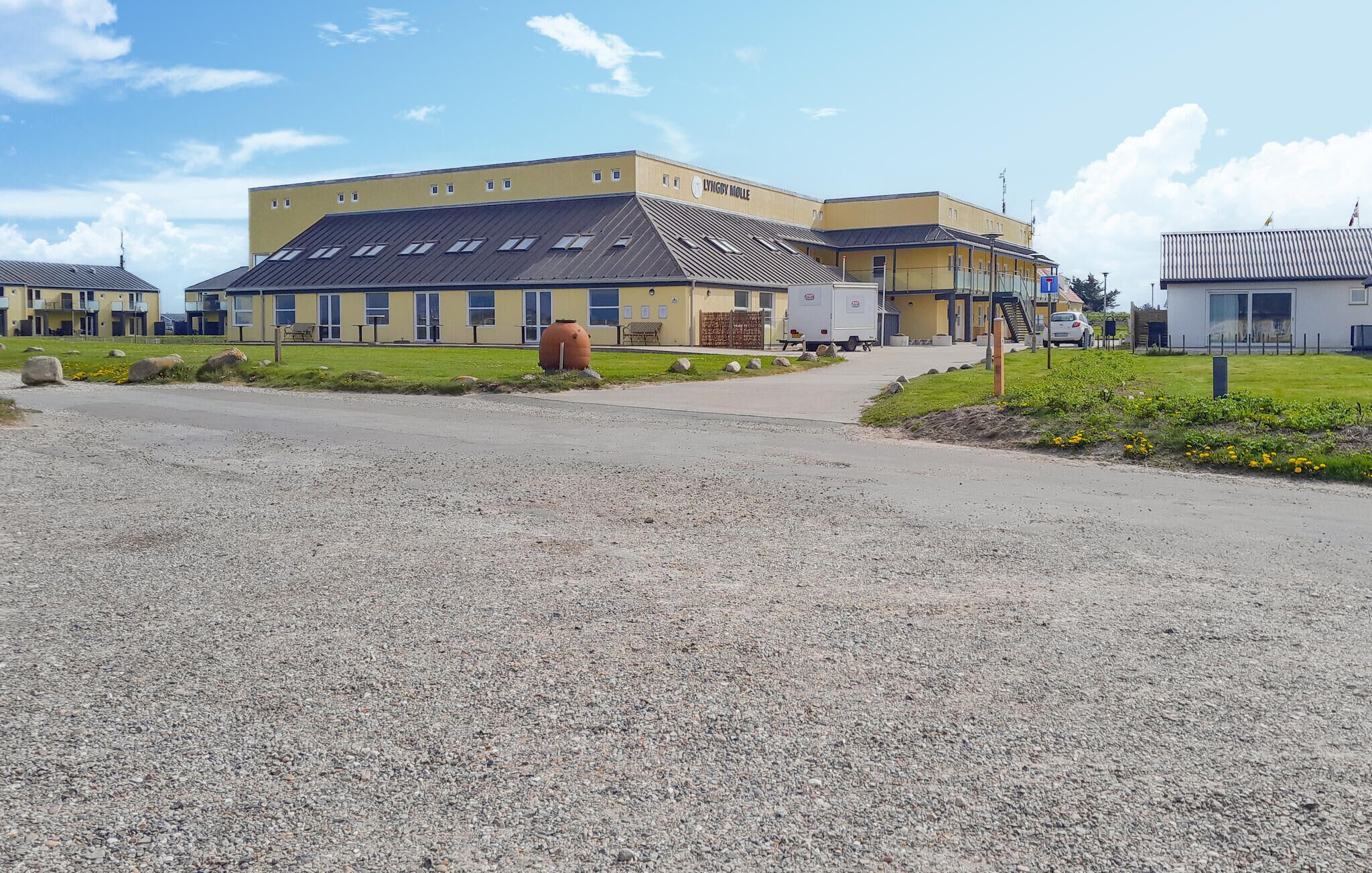 A building with a yellow facade and a grey roof, a gravel parking area, and a small garden with yellow flowers.