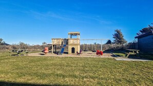 A playground with a wooden slide and swings, surrounded by a grassy yard and a building in the background.