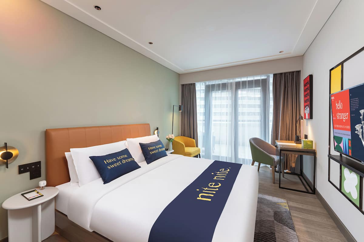 Habitación de hotel con una cama grande, una mesita de noche, una silla, un escritorio y un ventanal con cortinas.