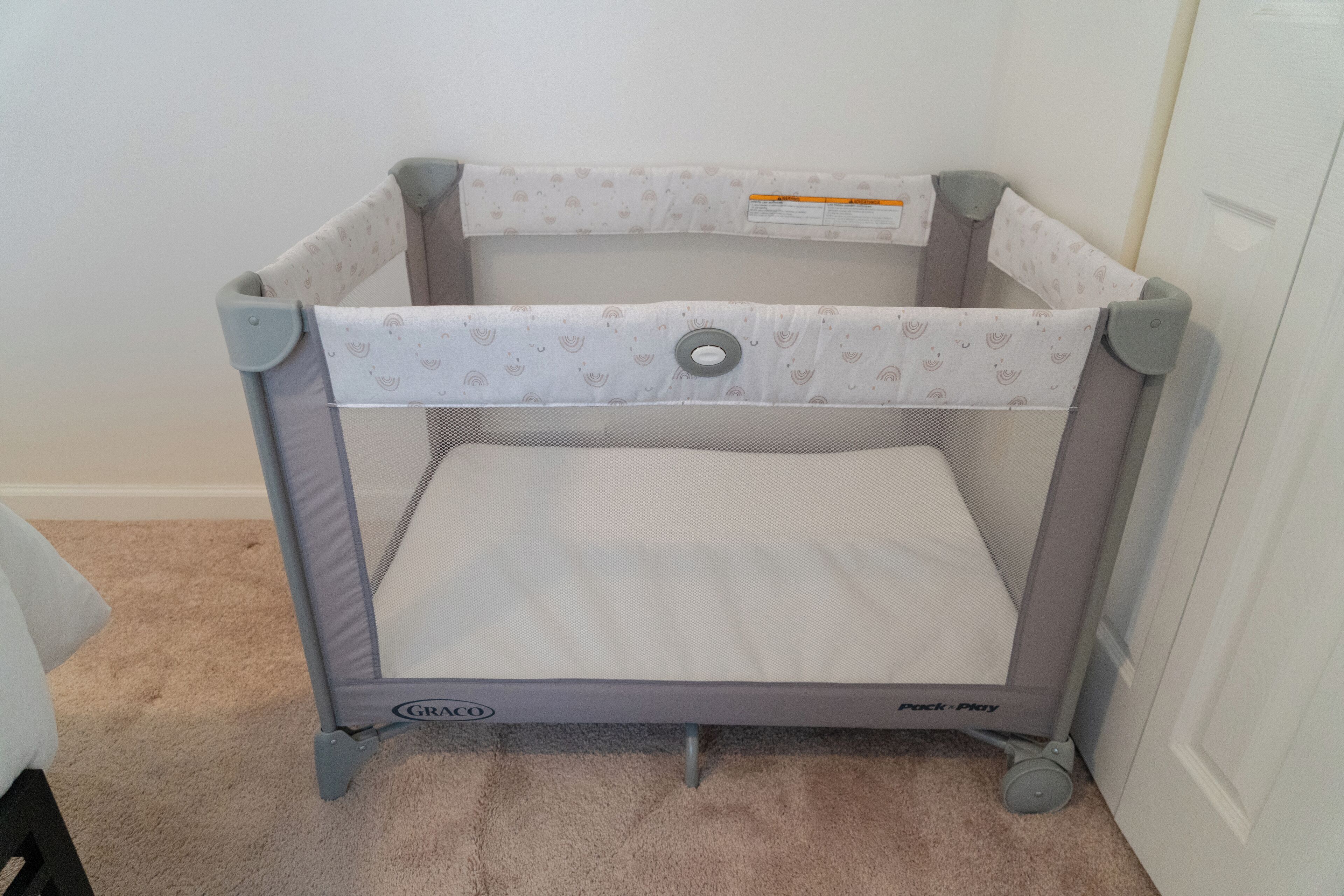 A portable crib with a mesh bottom and a padded top, featuring a safety strap and wheels for mobility.