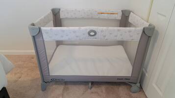 A portable crib with a mesh bottom and a padded top, featuring a safety strap and wheels for mobility.