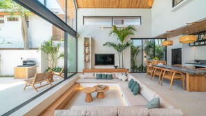 A modern living room with a large window, a wooden ceiling, a built-in TV, and a dining area with wooden furniture. - Villa Bayu by GORO (Seminyak)