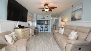 A spacious living room with a beige sofa, a coffee table, and a TV mounted on the wall. - Sea Oaks 103 - Oceanfront Complex in Garden City Beach (Garden City Beach)