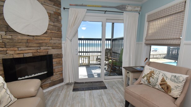 A living room with a stone fireplace, a beige sofa, a cushioned armchair, and a view of the beach through a sliding glass door. - Sea Oaks 103 - Oceanfront Complex in Garden City Beach (Garden City Beach)