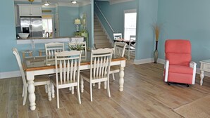 Un coin repas avec une table et des chaises en bois, une cuisine à l’arrière-plan et un escalier menant à un autre niveau.
