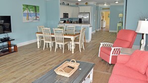 Un salon avec une table basse, un fauteuil rouge et un coin repas avec des chaises blanches, et une cuisine en arrière-plan.