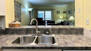 A kitchen with a double stainless steel sink, granite countertops, and a view into the living area. - Sea Oaks 102 - Oceanfront Complex in Garden City Beach (Garden City Beach)