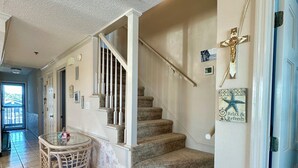 Un interior espacioso con una escalera que conduce a un nivel superior, un comedor con mesa y sillas, y una pared decorativa con una cruz y un cartel.