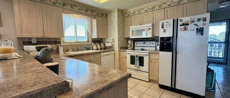 A modern kitchen with granite countertops, wooden cabinets, a built-in microwave, a refrigerator, and a window with blinds.