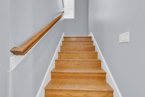 A staircase with wooden steps and a wooden handrail, leading to a landing with a white wall and a window with blinds.