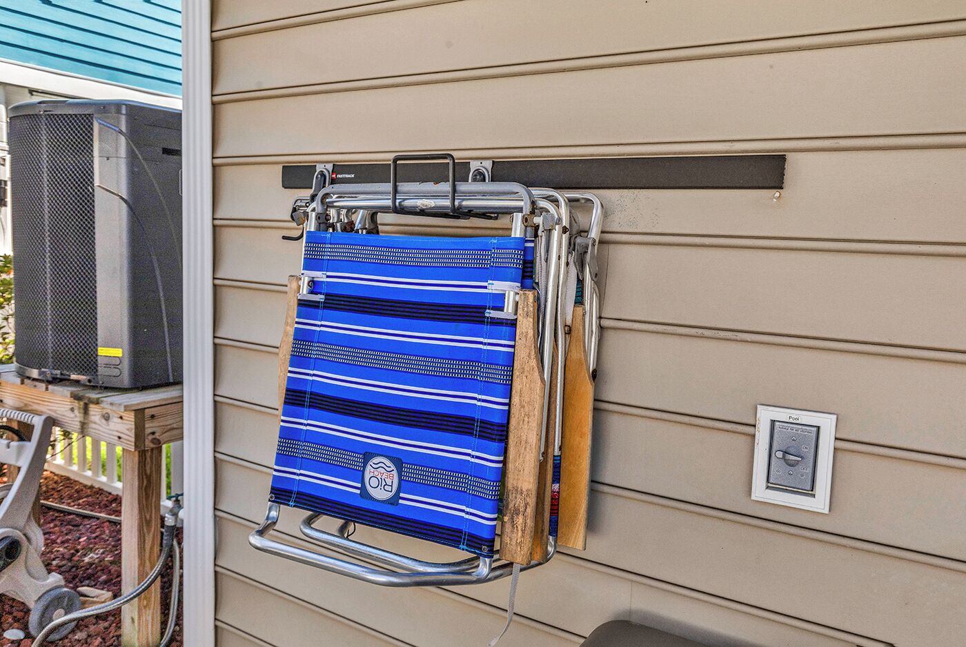 Uma toalha azul pendurada em um suporte metálico fixado em uma parede externa bege, com um painel de controle da piscina por perto.