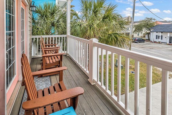 A wooden deck with Adirondack chairs and a blue cushion, surrounded by white railings and palm trees.
