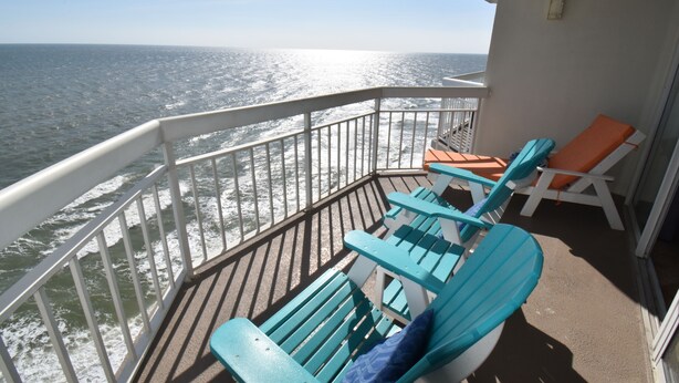 Ein Balkon mit türkisfarbenen Adirondack-Stühlen und einem orangefarbenen Stuhl, mit Blick auf den Ozean.